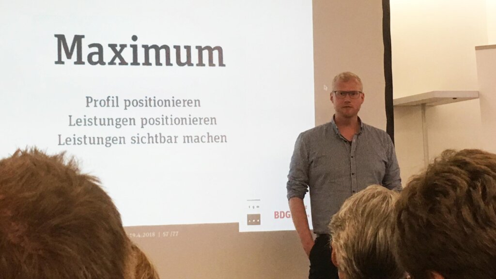 Ein Mann steht vor einem Publikum auf einer Bühne und hält einen Vortrag