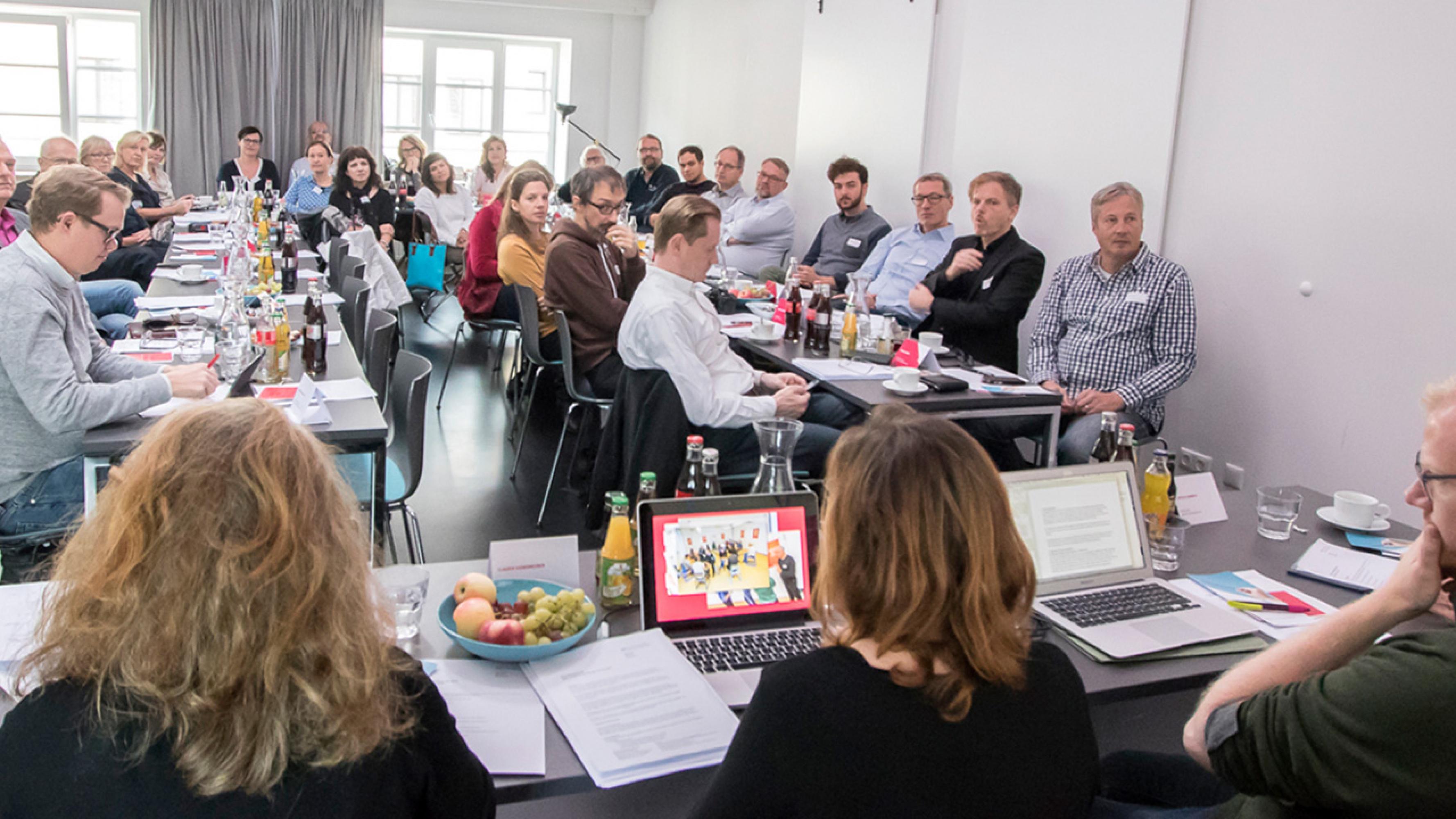 Etwa 30 Menschen sitzen in einem Meetingraum