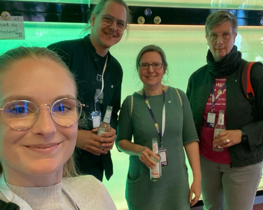 Selfie von Daniela mit Simon, Claudia und Birgit. Alle haben eine Wasserflasche in der Hand und ihre Konferenzbändchen um den Hals.